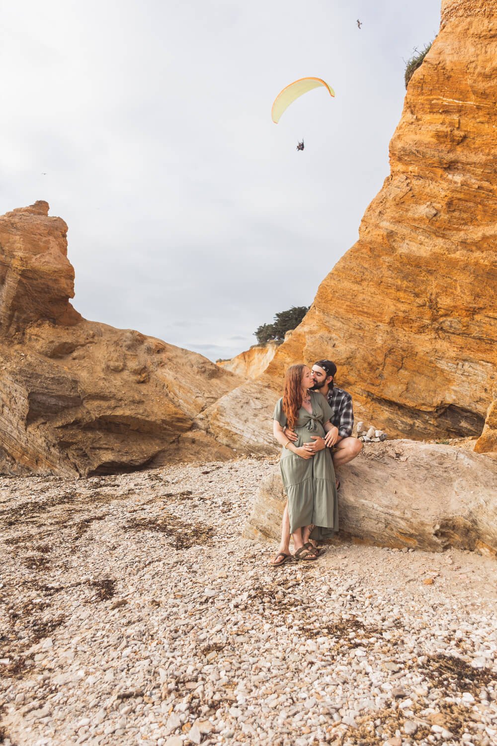 Séance photo grossesse en couple au bord d’une falaise Couple qui s’embrasse lors d’une séance photo grossesse en extérieur, avec une falaise et un parachutiste en arrière-plan